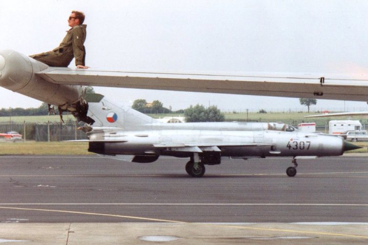 Pistonheads - In the image, a man is perched on the nose section of a fighter jet, which is parked on a runway. He seems to be inspecting the jet, possibly the pilot. Another jet is also visible in the image, further down the runway. The scene appears to be set during the day in an open area, likely an airport or airbase. There are also some trucks visible in the background, hinting at the operational nature of the location. The image has a grainy quality, typical of certain photographic styles or lighting conditions.