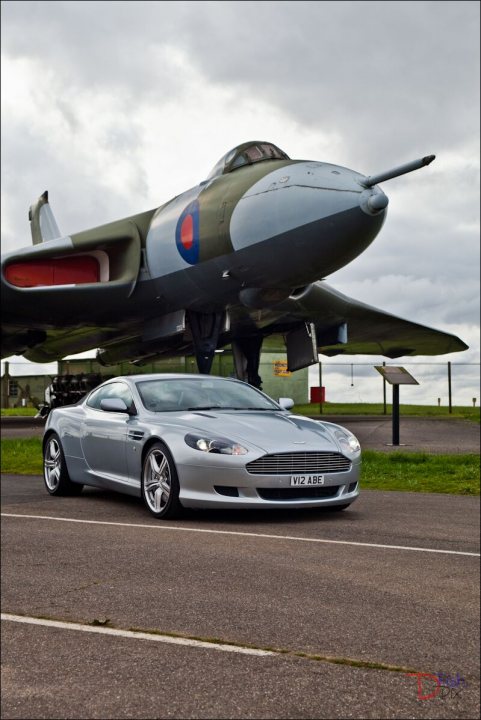 So what have you done with your Aston today? - Page 226 - Aston Martin - PistonHeads - This image features a silver sports car prominently positioned in front of a black, olivier green fighter jet with a camouflage pattern on the nose. The fighter plane, which has one of its wings extended horizontally, is partially parked on airstrip with the car adjacent to it. The sky above is cloudy, with dark and gloomy-looking clouds, suggesting an overcast day. The plane appears to be vintage, possibly a historic military aircraft. The sports car itself is likely a modern vehicle, with a distinctive design and smooth body.