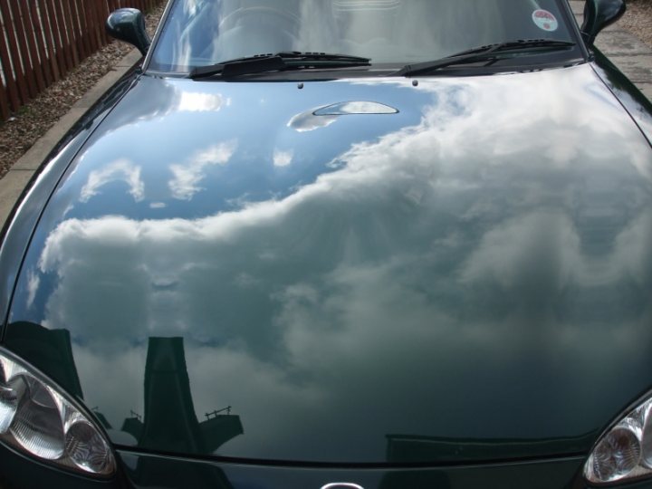 Shiney Pistonheads - The image shows a shiny, dark-colored car with its roof up, reflecting the sky. The car displays a logo indicating it's a convertible, common in the Mercedes brand. The sky above is a bright, blue one with a few scattered clouds, adding to the reflective quality of the car's surface. The car is parked in an outdoor setting that features a wooden fence in the background. The overall tone of the image is casual and it captures the luxurious and sun-dappled ambiance of a classic convertible.