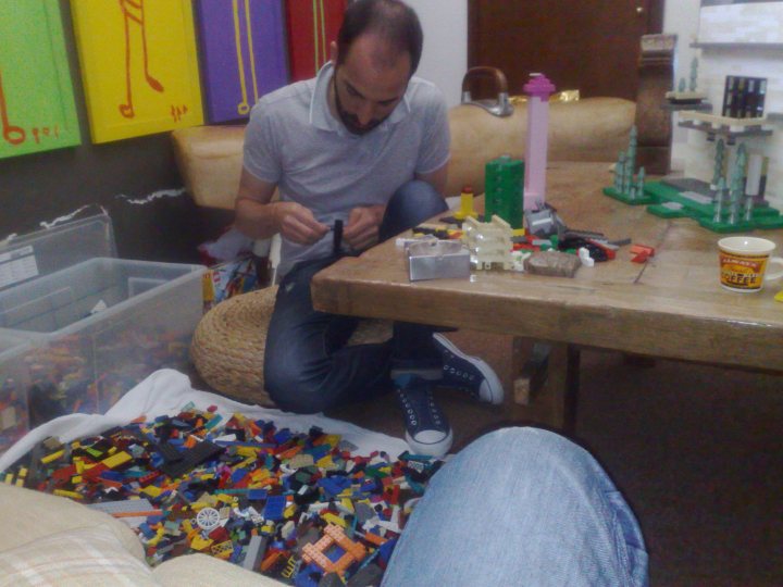 This image depicts a man sitting at a wooden table in a room filled with various items. The man appears to be engaged in a task, possibly assembling something, given that he is surrounded by toys and other small objects. His focus seems to be on these items, which are scattered across the table. The room has a casual, lived-in atmosphere, with a variety of colorful objects that add to its cluttered appearance. The man is wearing a gray shirt, and his expression suggests concentration.