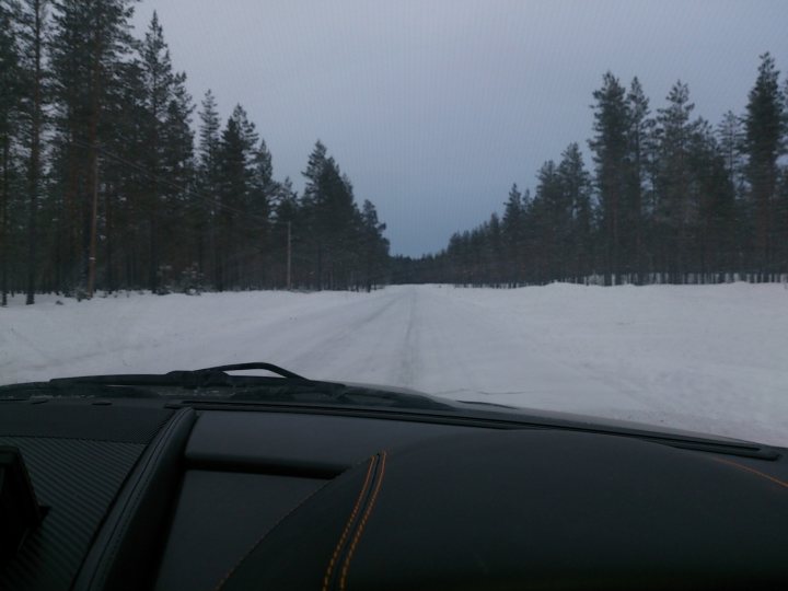 Aston Martin DB9 on Ice lake - Page 1 - Aston Martin - PistonHeads - The image shows a scene of a snowy wooded road on a clear day. The road is lined with evergreen trees on both sides, creating a symmetrical view. The snow-covered road is seen from the perspective of a vehicle's windshield, with the driver's side visible. The ground appears to be densely covered with fresh, untouched snow, reflecting the sunlight.