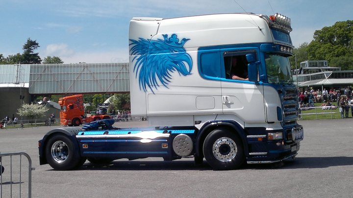 A large truck is parked in a parking lot - Pistonheads - The image features a large white truck that has a blue design on the front. This truck has a prominent yellow knight on the hood and a blue owl on the door. The cab of the truck is blue with the owl painted in more detail than the knight. A man can be seen in the cab on the left. The truck is on a road with grass and trees in the background, and several people are observing the truck from a distance.