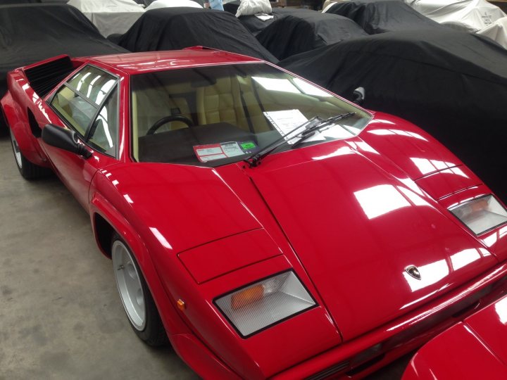 My special trip to buy a Countach in South Africa - Page 3 - Lamborghini Classics - PistonHeads - This is a photograph of a striking red convertible sports car. It appears to be located in a showroom or garage, with other red cars wrapped in plastic covers in the background. The vehicle has a sleek design with smooth curves, large wheels, and a prominent rear spoiler. The side badge and logos are visible, but I'm limiting the personalization aspect of my response. The car is not in use and seems to be polished and well-maintained.