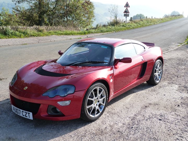 Canyon Red! - Page 1 - Elise/Exige/Europa/340R - PistonHeads - The image shows a red Ferrari sports car parked on the side of a road. The car features a prominent front grille with the Ferrari logo and two side vents. It has a sleek design with aerodynamic lines, and large headlights with blue accents. The car also has black side mirrors and five-spoke alloy wheels. The road itself appears to be a rural or suburban location, and there's a glimpse of a verdant landscape on the horizon. The time of day appears to be morning or late afternoon due to the soft lighting.