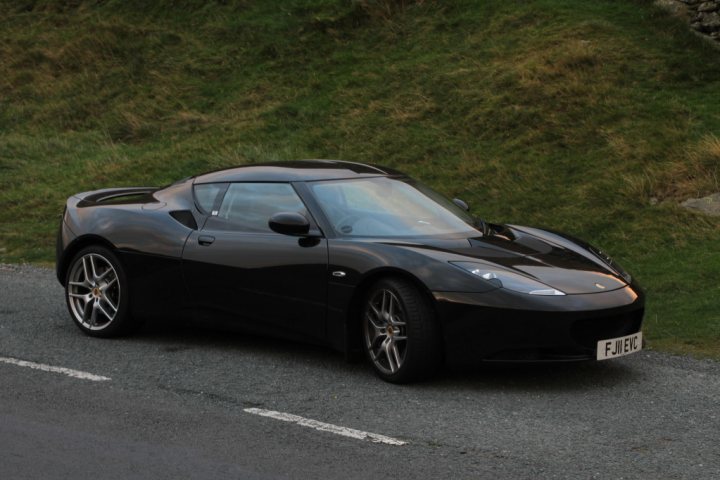 black evora spotted - Page 1 - Evora - PistonHeads - The image shows a sleek, black sports car parked on a roadway. The car has a low-to-the-ground design and features two doors. One noticeable feature is the regenerative braking system, detailed on the side of the vehicle near the front wheel. The road appears to be a country or suburban street, with lush green grass on either side, indicating a calm, residential environment.