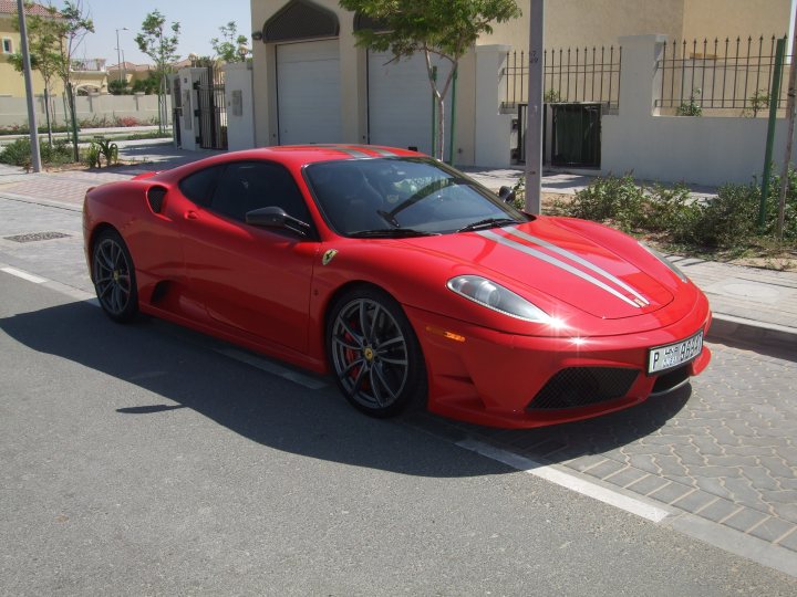 My First Ferrari...and its a Scud! - Page 1 - Ferrari V8 - PistonHeads - The image captures a vibrant scene on a city street. Dominating the foreground is a sleek red Ferrari sports car, its polished body gleaming under the sun. Its black side mirrors reflect the surrounding environment, including a white car parked nearby. The Ferrari is situated on a sidewalk adjacent to a street, hinting at a bustling urban setting. In the slightly blurred background, elements of city architecture can be seen, including a building with a green fence and a tree offering a touch of nature amidst the urban jungle. The photograph appears to be taken from a distance, capturing the Ferrari and its surroundings without foreground obstruction.