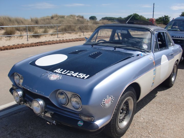 Scimitar Pistonheads Coupe - The image features a classic black and silver Allu245H sports car parked on the right side of the frame. The car is branded with a large circular logo on the hood and additional logos on the doors. The vehicle's design includes a unique European license plate symbol, suggesting that this car is of European origin. The background reveals a scenic location, possibly near a beach or a rural area, with the car on a dirt road next to a metal railing.