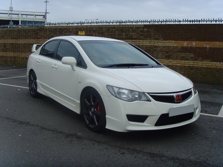 Honda Pistonheads Fdr - The image shows a white two-door sedan parked in a parking lot against a wall with a fence on top. The vehicle has a black roof and black rims, contrasting with its white body. It features a visible logo on the grille and a rear brush on the back bumper. The photograph has a grainy texture and a slight angle, capturing the front passenger side of the car. The bridge in the background suggests an urban setting.