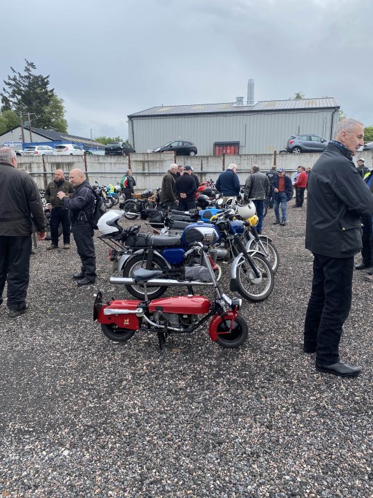 A picture a day...biker banter (Vol 6) - Page 112 - Biker Banter - PistonHeads UK - The image shows an outdoor scene where a group of people is gathered around various motorcycles parked on a gravel surface. There are at least five motorcycles visible, each with different colors and designs. Some motorcycles have sidecars attached to them. The people present seem engaged in conversation or inspection of the bikes. In the background, there's a fence and some buildings, suggesting that this event is taking place in an urban or semi-urban setting. The presence of a tie indicates that some attendees are dressed formally. The atmosphere appears to be casual yet social, with a sense of camaraderie among the motorcycle enthusiasts.