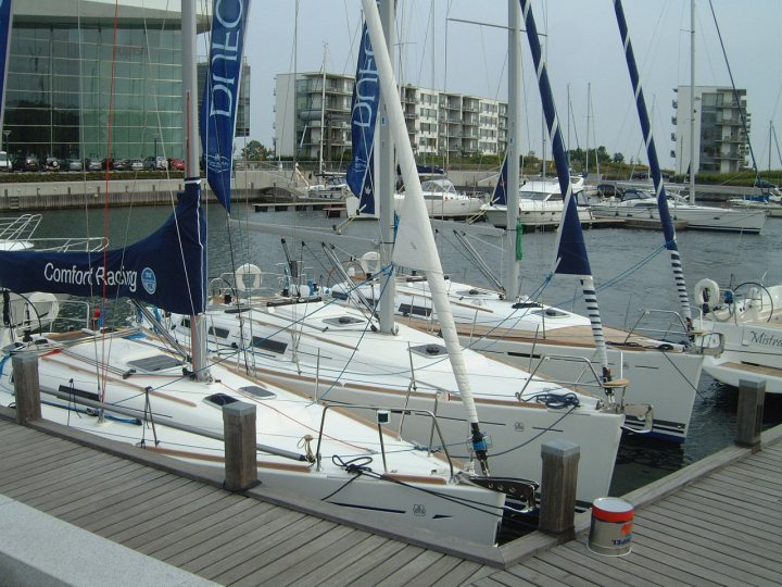 The image presents a tranquil scene of a marina filled with moored sailboats. There are several boats parked side by side, their white hulls gleaming in the water, suggesting they're ready for a day out on the sea. Each boat is adorned with blue sails loosely mounted on tall masts, lightly fluttering in a gentle breeze. The dock, which appears to be made of wood, juxtaposes nicely with the steel structures of the residential buildings visible in the background. The calm water of the marina is dotted with a few buoys, adding to the overall serene ambiance of the nautical setting.