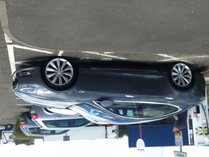 Friend's Tesla S  - Page 1 - EV and Alternative Fuels - PistonHeads - This image provides a unique aerial view of a parking lot, where several cars are parked solely on the front wheels. A silver-colored sedan is situated in the foreground, with its front wheels resting on the curb of the parking space. Behind it, another car is parked with its front wheels on the street, creating a diagonal tilt that adds to the unusual perspective of the photo. In the background, gas pumps are visible, indicating that this scene is taking place at a gas station. The image is taken from a high angle, emphasizing the unusual elevation of the vehicles.