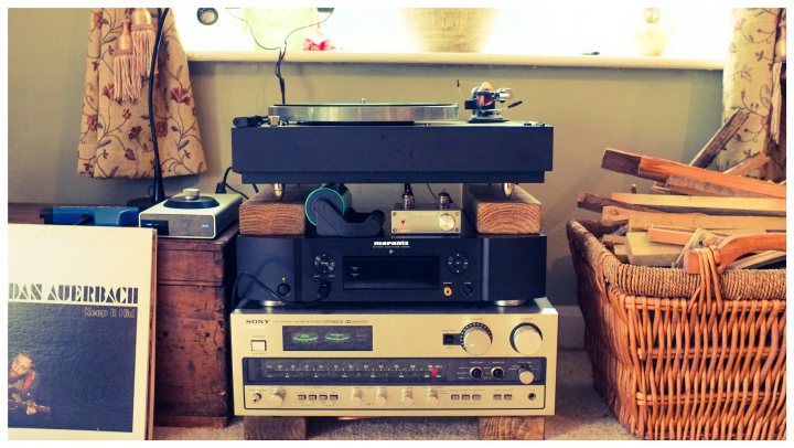 What’s your Hi-Fi set up? spec and pictures please  - Page 20 - Home Cinema & Hi-Fi - PistonHeads - The image depicts a table set up for DJing or recording. There's an old-fashioned record player at the center, with a stack of records next to it. Above the record player is another machine that appears to be part of a radio or sound system setup. To the left of the record player, there are a couple of speakers, possibly part of the sound system. There's also a small pile of books, which might contain music scores or other related materials. The table is set against a wall and seems to be in an indoor setting, as suggested by the presence of what looks like a window or curtain. Overall, it's a setup that suggests the creation or enjoyment of music.