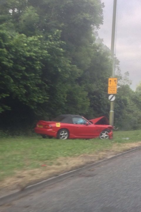 I wonder what happened here then? - Page 1 - Thames Valley & Surrey - PistonHeads - The image depicts a scene on a road where a red convertible sports car has been involved in a crash, having veered off the road and collided with a pole. The car is now rendered inoperable, resting precariously against the pole. The surrounding environment is lush with green trees, indicating that the crash has occurred in a suburban or semi-rural setting. Safety cones and a yellow sign with black symbols, possibly indicating the presence of a construction or slippery road area, are visible near the car crash site. During the day, the natural light illuminates the scene, casting shadows and highlighting the road and the aftermath of the accident.