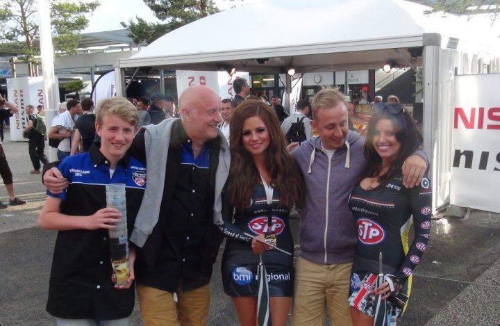 RAM at tunnel for Lemans trip - Page 1 - Vipers - PistonHeads - The image shows a group of five people, posing for a photo. They are outdoors on a sidewalk, standing under an open-air gazebo. On the right side of the photo, there is an advertisement, which seems to be for a car, prominently displayed. The individuals in the photo appear to be participating in an event, as indicated by the photo backdrop with the number 75 visible in the background.