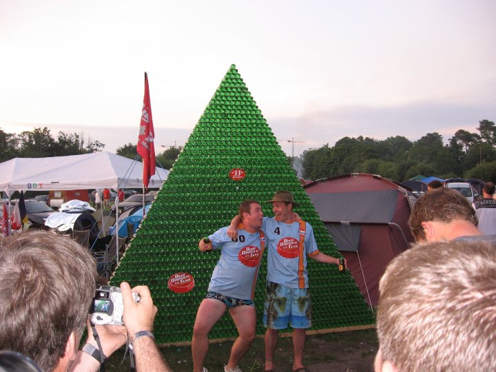 Mountain Beer Pistonheads - The image features a vibrant and cheerful scene at what appears to be a festive or outdoor gathering. In the center, two individuals are playfully pretending to push the number 38 up a steep, humongous green mountain made entirely of stacked plastic bottles. Other attendees are scattered around the area, facing the central scene, some capturing the moment with their cameras and cell phones. The background reveals a cloudy day and the outlines of tents, possibly housing the event's attendees. The grassy field where the action is taking place suggests a park or open outdoor space.