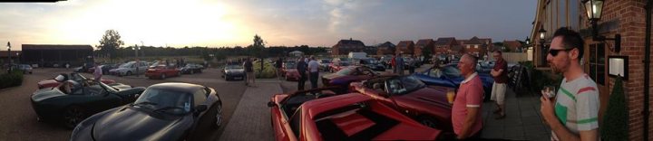 TVR + Guests Berkshire July meet - Page 3 - Thames Valley & Surrey - PistonHeads - The image is captured from a person standing on a street. The setting includes a variety of parked cars on the side of the road. It appears to be around sunset since the sky is showing hues of the sun going down. The cars include both vintage and modern models, suggesting a diverse crowd or event. The image is a bit blurry, which might be due to the effect of the evening light or the camera's focus.