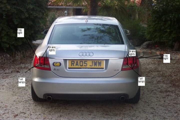 Law Pistonheads Foglamps - The image shows the rear view of a silver Audi car parked on a gravel surface. A red brake light is prominently visible on the upper section of the rear bumper, slightly to the left of the center. The chrome badge on the rear bumper has the following text: GGB-AUST, RAC50J, and AUA-SYDON. Two black inspection stickers can be seen above the license plate, which reads: RAO5JWM, the numbers white against the yellow background. To the right of the license plate, the tail light is labeled as "Tall emps."