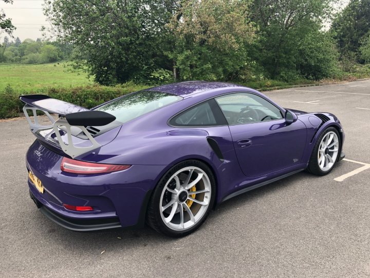 Porsche 991 GT3RS Ultra Violet  - Page 1 - Readers' Cars - PistonHeads - This image captures a striking purple Porsche sports car parked in a lot. The car is positioned on the left side of the frame, with its front facing the viewer. It's equipped with a large rear wing and gold wheels, adding to its sporty aesthetic. The vehicle appears to be in pristine condition, suggesting it may be new or well-maintained. In the background, there is a serene landscape featuring greenery and trees under a clear sky, providing a peaceful backdrop for the car.