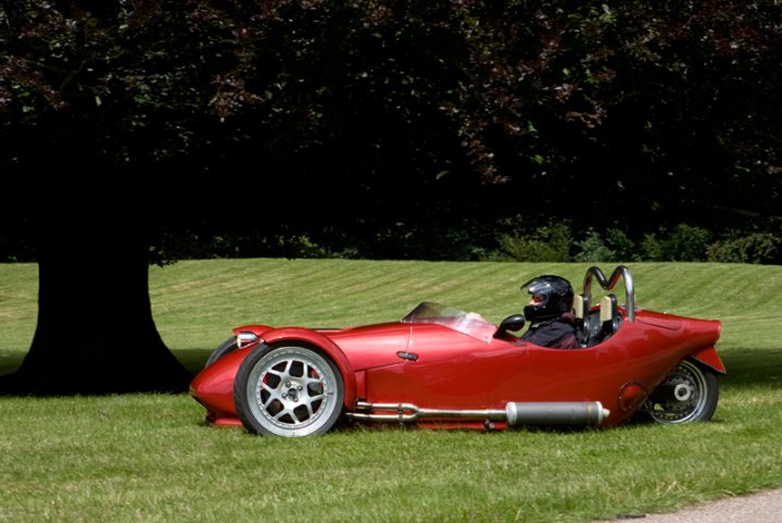 Pistonheads Motors Manor - In the heart of a serene green park, a person is seated on a futuristic-looking red three-wheeler vehicle. The vehicle has a distinctive design with a strong body and a single large wheel at the front. The person, wearing a helmet, takes the driver's seat, positioned centrally on the vehicle, ready to navigate this three-wheeled marvel through the lush greenery. The sky overhead is overcast, adding a dramatic backdrop to this tranquil scene. The park itself is expansive, with a single large tree standing prominently near the driver's vehicle, adding depth and a touch of wilderness to the scene.