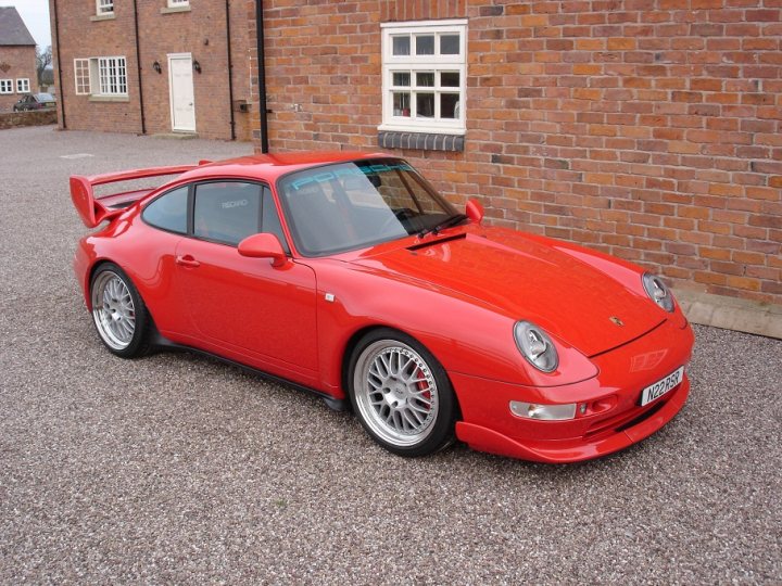 Slippydiff Pistonheads - The image shows a vibrant red Porsche 911, parked on a gravel driveway. The car's long front nose and extended rear give a sense of its powerful performance capabilities. It features a classic white roof and a black fabric soft top. The setting is a gravel surface adjacent to a red brick building with a white window frame. The overall scene suggests a residential or possibly a boutique garage setting.