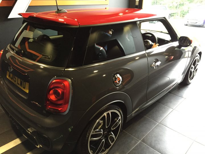 New arrival - Manual F56 JCW - Page 1 - New MINIs - PistonHeads - This is a photograph of a compact sports car parked indoors, possibly in a showroom or on a car dealership floor. The car's color scheme includes a red roof and a black or dark grey body, with an orange badge center top, perhaps indicating a specific model from the Mini brand. The car has a five-door hatchback configuration, two black doors visible in the side profile. The design of the vehicle suggests it's a recent model, with the expected roundel of the Mini symbol and the distinctive wheel design. The photograph captures the vehicle from a rear three-quarter angle, and it is viewed against a neutral grey background with artificial lighting, given the indoor setting.