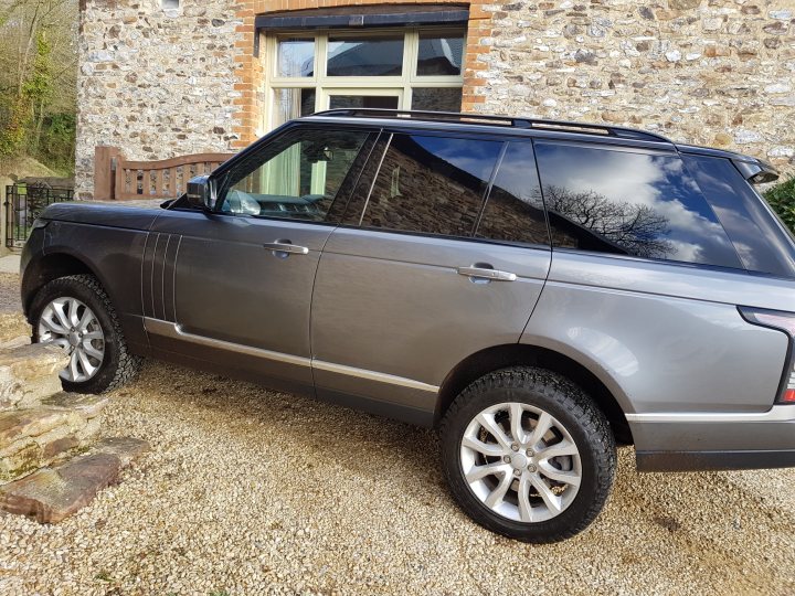 L405 proper off road tyres? - Page 1 - Land Rover - PistonHeads - The image shows a dark gray Land Rover parked on a gravel driveway. The vehicle is a four-door SUV with a visible spare tire mounted on the rear door. A set of stairs and a stone building can be seen in the background, suggesting a rural or semi-rural setting. The skies are overcast, and no people are visible in the image. The style of the image appears to be a standard, non-professional photograph taken during the day.