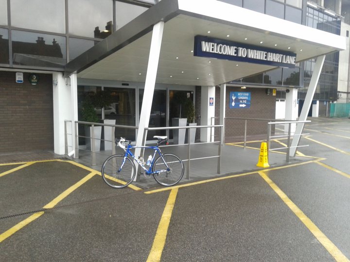 The "Photos From Today's Ride" thread... - Page 150 - Pedal Powered - PistonHeads - The image shows the entrance to a building that is being used for accommodations, indicated by the "Welcome to Whitehall" sign. There is a light blue bicycle parked outside the entrance, which is secured by a chain. A pathway leads up to the main doors, which are currently open, revealing an interior that seems to be a lobby or reception area with a few potted plants. The ground is covered with a pattern of yellow and white lines denoting parking spaces.