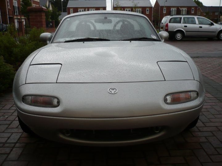 Pistonheads Bodyshop Booked - The image shows a gray sports car parked on a brick driveway. The car's body appears to have some raindrops on it, suggesting it may have been raining recently. In the background, another vehicle is also visible, parked next to the sports car. Beyond the driveway, residential houses are visible, indicating that the cars are parked in a residential neighborhood.