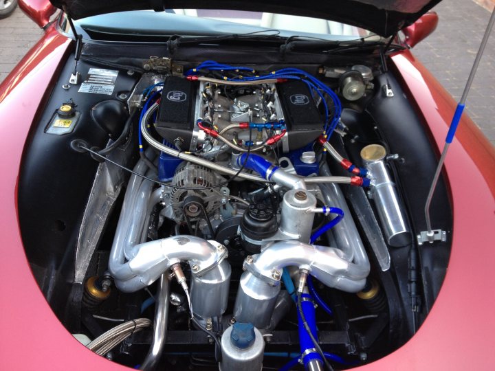 ACT manifolds  - Page 1 - Chimaera - PistonHeads - This image provides a close-up view of the undercarriage of a vintage sports car. The engine area is visible, showcasing intricate mechanical components. The car's interior has red-hued paneling, contrasting with the cool blue of the exhaust system. The open bonnet reveals the hood mounts, a common feature in sports cars. The engine bay is compact but highly detailed, indicative of the vehicle's high performance capabilities.