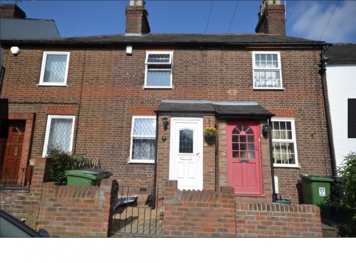 Determining mid-terrace wall thickness? - Page 1 - Homes, Gardens and DIY - PistonHeads - The image depicts a red brick house with white windows and white trim. The house has a symmetrical design with two entrance doors. One door is painted red and the other is white, both adorned with decorative glass pane windows. Above the red door, there's a small balcony with a flower pot that adds a touch of color to the scene. To the left of the house, we see a green metal trash can sitting next to a steel rail, suggesting a residential street scene. The sky is blue, indicating a clear day.