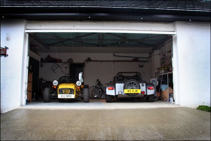 Not enough pictures on this forum - Page 17 - Caterham - PistonHeads - The image shows an indoor mechanic's garage with two vintage cars parked inside. The vehicle on the left is a bright yellow open-top car, while the one on the right is a black and silver open-top car. Behind the cars, there is a workbench and various tools and equipment. A garage door with reinforced, corrugated metal panels fills the background.