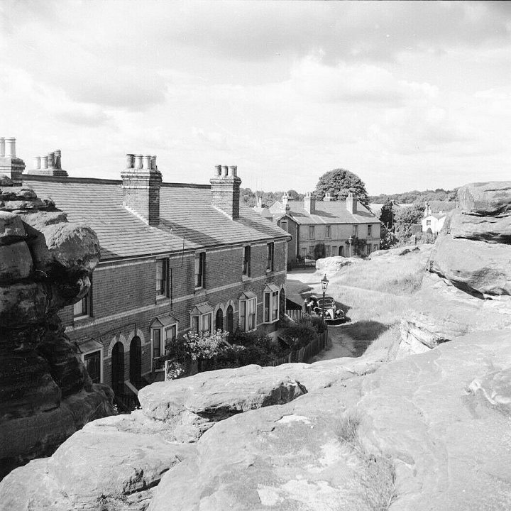 Identify the location from old photos - Page 1 - Photography & Video - PistonHeads - The image is a black and white photograph of an old-fashioned cityscape. It features a street lined with buildings, some of which have chimneys, suggesting the presence of fireplaces. On either side of the road, there are rocky cliffs that add a rugged charm to the scene. A few cars can be seen parked along the street, and a solitary figure is visible in the background. The overall atmosphere of the image suggests a peaceful town nestled amidst nature.