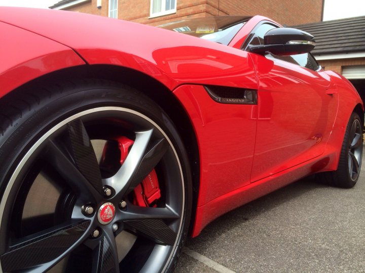 Whats the best colour for a F-type coupe - Page 1 - Jaguar - PistonHeads - The image shows a vibrant red sports car with distinctive black rims and spokes, positioned outdoors. The car is parked on a driveway, with the perspective focused on the front tire. The design of the rims, with a visible pattern and cutouts, suggests the car could be a luxury or high-performance vehicle. The surface of the rims reflects some spots, indicative of a polished or well-maintained exterior. In the background, there is a structure with a brick wall, and part of a garage door is visible, suggesting the location of this scene is a residential area.