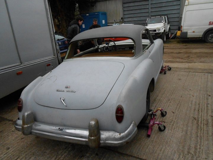 Nash Healey - Page 1 - Classic Cars and Yesterday's Heroes - PistonHeads - The image shows an old, grey convertible car parked on a concrete surface, with one door open revealing the interior. The car appears to be a vintage model, potentially a production model due to the "Lincoln" logotype visible on the rear of the car. The setting seems to be a parking area, possibly in an industrial or commercial zone, as indicated by the presence of multiple trucks and other vehicles in the background. The car's silver exterior suggests that it has been well-maintained, and the sunlight reflecting on it indicates that the photo was taken during the day in good weather conditions.