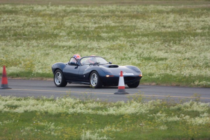 Ajec Racing Trackday at Gloucester Airport - Page 1 - South West - PistonHeads - The image captures a scene of a vintage sports car embracing the speed of a highway. The car, with its sleek design and open-top architecture, is currently driving away from a line of caution cones. The backdrop is a lush, green field filled with flowers, contrasting with the vehicle's metallic sheen. As it races along, the driver in the car is donning a helmet, suggesting the thrill and danger of the adventure.