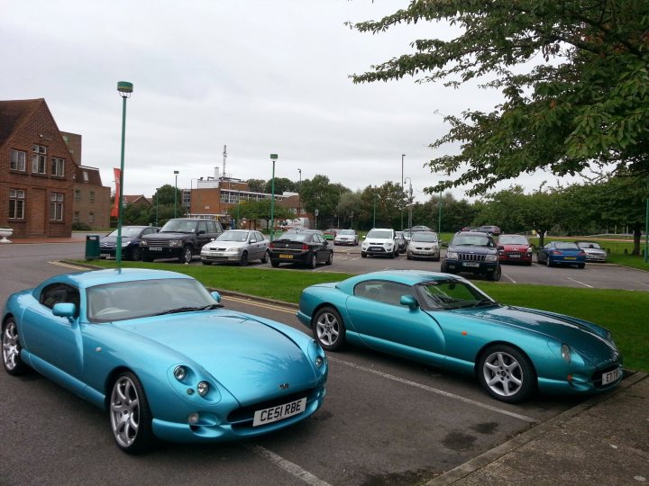 Differences between Mk1 and Mk2 - Page 1 - Cerbera - PistonHeads - The image features a parking lot where two striking teal sports cars are parked. The cars are parked in adjacent spaces, one closer to the camera and the other farther away, creating a sense of depth in the image. Parked next to these sports cars, and filling up a larger part of the image, are several ordinary cars. The lot is well-maintained, with clearly marked spaces, and there's a green and white building visible in the background. Despite the presence of multiple vehicles, the focus of the image remains on the teal sports cars.