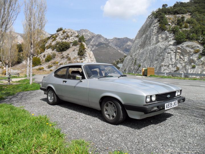 Pistonheads - The image captures a moment of solitude on a windswept mountain pass. Dominating the center of the frame, a classic British sports car paints a picture of nostalgia. The car, parked on a rocky road, is older but still maintains her sleek and strong silhouette. The landscape around the car is rugged and unspoiled, with majestic mountains rising in the distance. The sky above is a brilliant shade of blue, dotted with a few fluffy clouds, adding to the serene beauty of the scene.