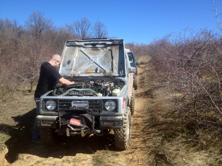 Bit of off-roading, and a bit of camping. - Page 1 - Off Road - PistonHeads - In the image, a man is working on the engine of a gray pickup truck. The truck is parked on a dirt road, and the man appears focused on the task at hand. The area around the truck is described as "bare and brushy," suggesting a lack of vegetation and an overgrown but not completely coverd road. The truck is in the process of hitting something on the road, as indicated by the implied contact with an unseen object. There is a small white line in the road, which could be a boundary marker or a route indicator. The truck's license plate reads "pro," but due to its content and privacy, it cannot be provided. The image captures a moment of human intervention with machinery, set against a natural landscape.