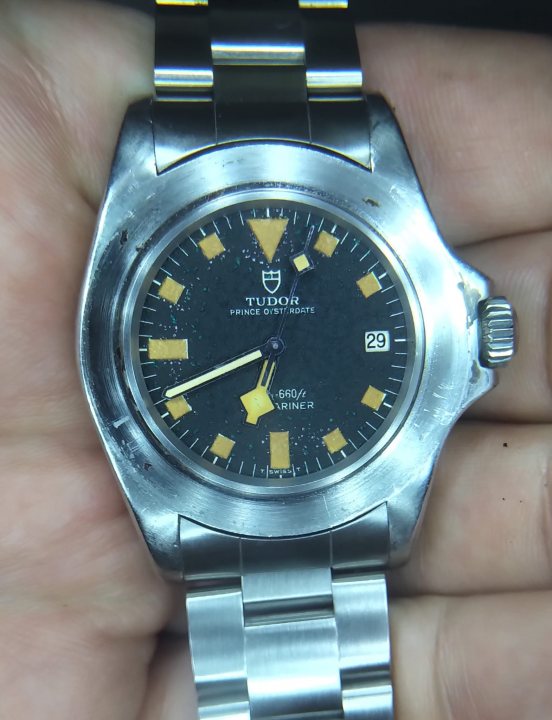 The image shows a person holding a wristwatch in a hand. The watch has a stainless steel band and a silver case. The watch face is black and features yellow numerals, indicating it's a Tudor model. On the face, the word "PRINCE" is written in white.