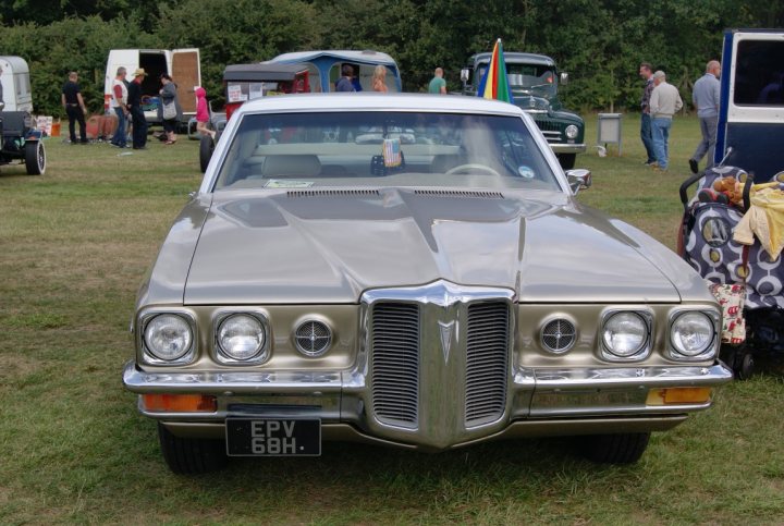 Anyone going to the NASC 40th Nats at Trinity Park? - Page 1 - East Anglia - PistonHeads - The image captures a scene at an antique car show. A vintage Pontiac GTO, characterized by its shiny silver hood and front grille with a chrome center, is parked on the grass at the event. Surrounding the car, there is a crowd of people who appear to be attendees of the show. In the background, there are several other vintage vehicles, and the presence of trees and a flag indicates that the event is taking place outdoors. A few individuals can be seen carrying suitcases, possibly containing items for sale or display at the show.