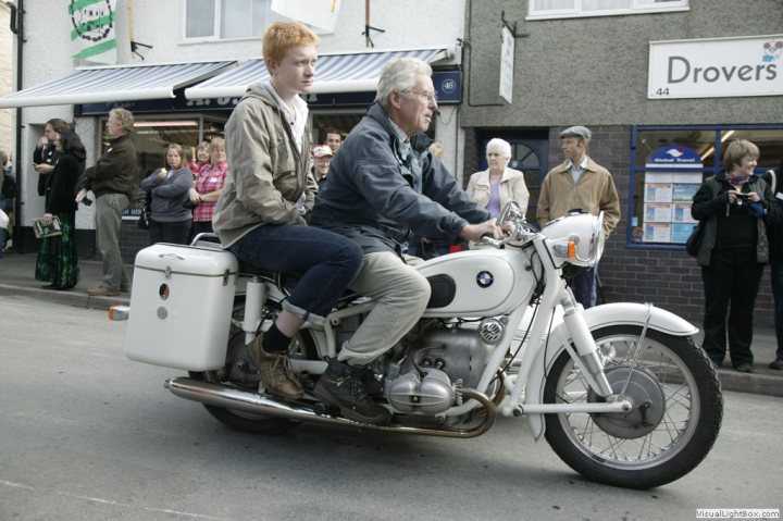 Classic tractors - Page 3 - Classic Cars and Yesterday's Heroes - PistonHeads - The image captures a dynamic street scene where a man and a boy are riding a large white motorcycle. The man, possibly the driver, is seated towards the back of the motorcycle, while the boy is perched on the front. Both are clad in casual attire and seem to be enjoying their ride. The motorcycle is being driven down a street lined with various small shops and cafes, adding a sense of urban charm to the scene. Besides the two on the motorcycle, there are several other individuals scattered in the background, contributing to the lively street atmosphere.