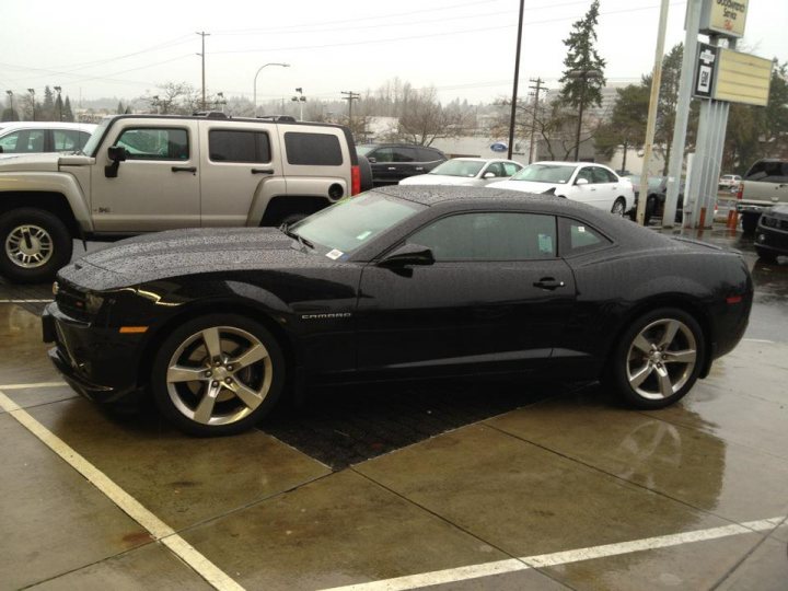 New car purchase! - Page 1 - Yank Motors - PistonHeads - In the image, a sleek black convertible is parked in a parking lot on a rainy day. The car is positioned in a space, surrounded by several other cars and trucks. The parking lot reflects a somewhat busy environment, and the wet pavement and contrasting drop-top roof of the convertible in the spotlight are the focal points of the photo. The photograph seems to be taken in a real-life setting, possibly during dusk, as suggested by the soft lighting.