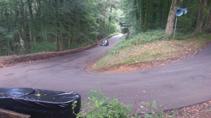 Pistonheads - The image is a photograph depicting a curved rural road. The perspective of the photo is from someone standing beside the road, looking down it toward the horizon. A black car is visible on the right side of the road, driving away from the viewer's position. The surrounding landscape includes trees and grass, suggesting a natural or suburban environment. There is no text in the image, and it has a candid quality, capturing a moment rather than staging a scene.