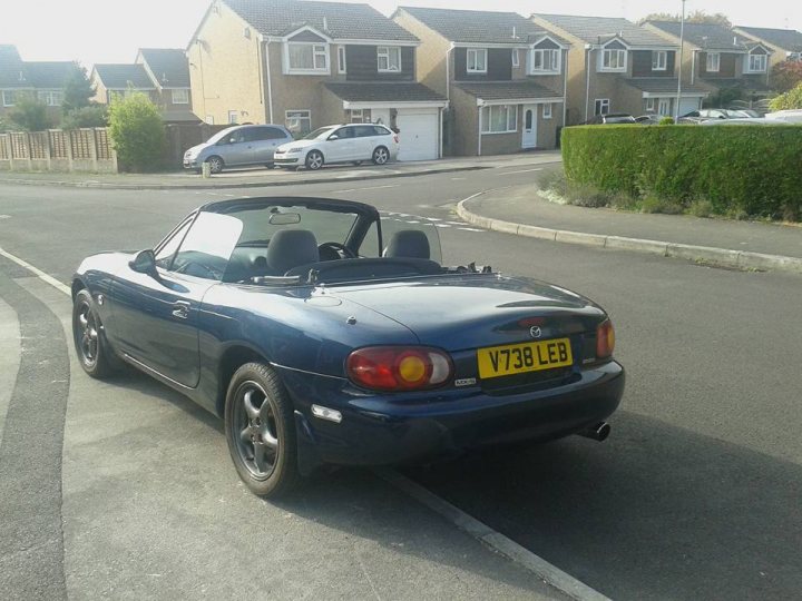 Got myself an MX-5 at last!! - Page 1 - Mazda MX5/Eunos/Miata - PistonHeads - The image depicts a residential setting with a curved roadway. Parked on the left side of the image is a blue car with a convertible top. The car is on the kerb, adjacent to a sidewalk, and is facing the opposite direction of the majority of the image. There are also two parked cars to the rear of the blue car. The cars are stationary, suggesting a quiet or residential area. The scene appears to be set under clear skies, possibly during a sunny day.