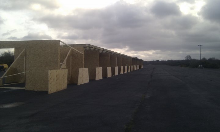 Pistonheads - The image features a row of several beige plywood panels neatly aligned next to each other beside a paved surface. The plywood panels are in varying lengths and are part of a construction site. The skies overhead are cloudy, suggesting an overcast or possibly late afternoon setting. The road leading up to the panels is empty and is bordered by a grassy area on one side. There is no visible text or branding in the image. The style of the image is a straightforward, unembellished photograph, which focuses on capturing the scene of the plywood panels and the surrounding environment.