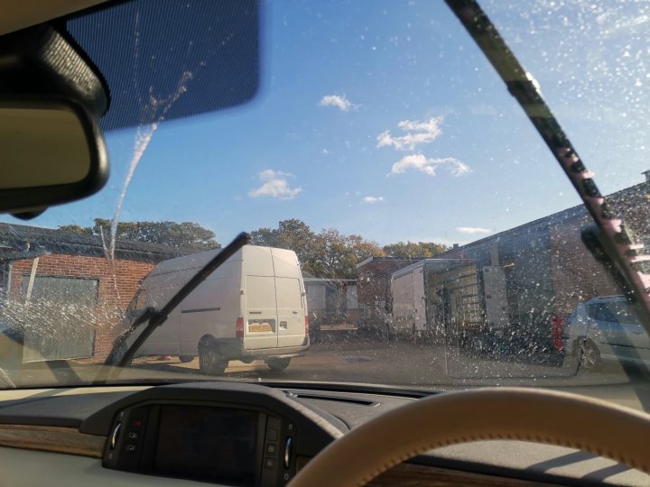 The image shows the interior of a car from the perspective of a passenger in the front seat. It appears to be taken during the day, as indicated by the bright lighting and clear visibility. Through the window, one can see a building with trucks parked outside, suggesting an industrial or commercial area. The sky is visible, indicating fair weather conditions. There are no people present in the image; however, there's a steering wheel on the left side of the vehicle, suggesting it's likely designed for driving on the right side of the road.