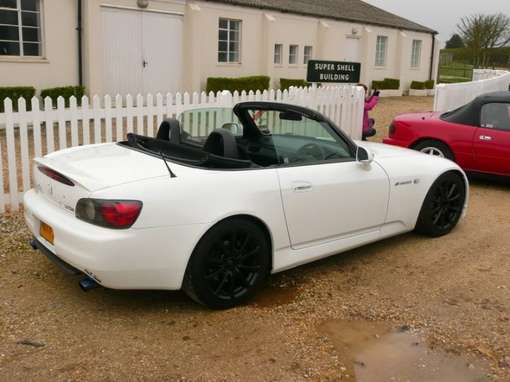 Pistonheads - The image showcases a pristine white sports car parked on a gravel surface in the foreground. The car features a sloped white roof and black tire rims, indicating a modern and sleek design. In the background, there are two other cars, one red and the other white, parked behind a line of white picket fence. The setting appears to be a residential area, with a house and a hedge visible. The car has a large spoiler, suggesting it might be used for performance or racing.