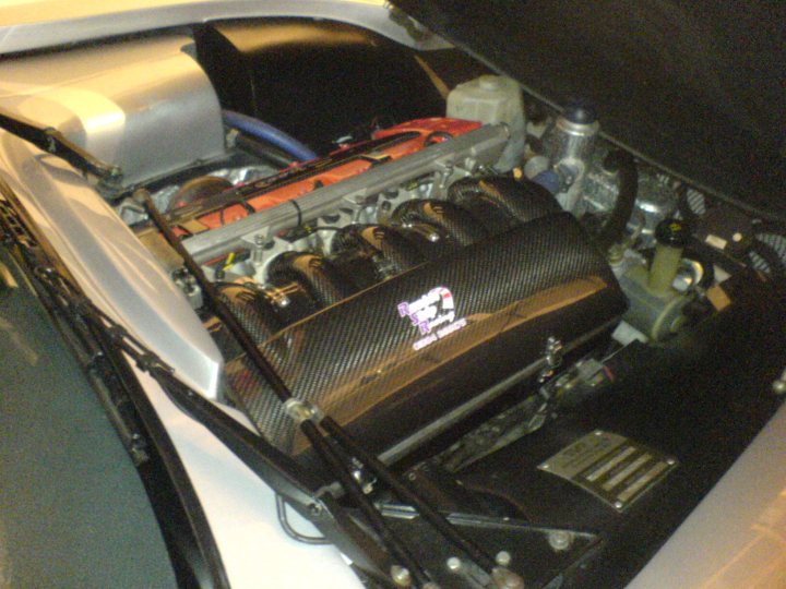 Some pictures of my Tam - Page 1 - Tamora, T350 & Sagaris - PistonHeads - The image shows the engine compartment of the back end of a light-colored car, with the hood lifted. The engine, a large V8 with a metallic finish, occupies the majority of the image. The uniform carbon cover is labeled "Kobe" with a checkered border. Various components and connections are visible around the engine, but the specific details are not clearly discernable. There is also an oil filter visible, indicating preventive maintenance. The lighting conditions seem to be captured in a garage or workshop, as indicated by the blurred background.