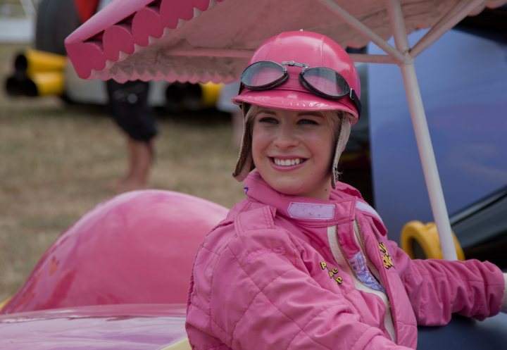 Pistonheads Goodwood Hunnies - In the center of the image, a young woman stands confidently, donned in a vibrant pink racing suit with a helmet. She is laughing, her smile bright against the blue sky that serves as her backdrop. Her pink helmet matches her outfit, and she holds a pink parasol in her right hand, adding a playful touch to her ensemble. The background reveals a tracking device lying on the ground, hinting at a racing or sports event.