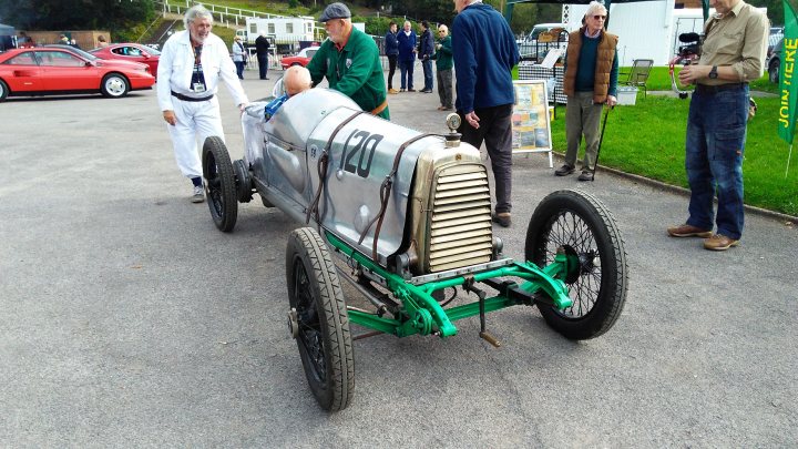 The rarest car you have photographed/seen - Page 12 - General Gassing - PistonHeads - The image shows an outdoor scene where a vintage car is being displayed. This small, single-seater vehicle with its engine exposed has a classic design, painted in white and green with black wheels. A man stands next to the car, inspecting it closely. His position suggests he might be the owner or a mechanic admiring the craftsmanship. In the background, there are a few people scattered around, possibly onlookers. The setting appears to be a street or a parking lot with some greenery visible, adding to the relaxed and casual atmosphere of the event.